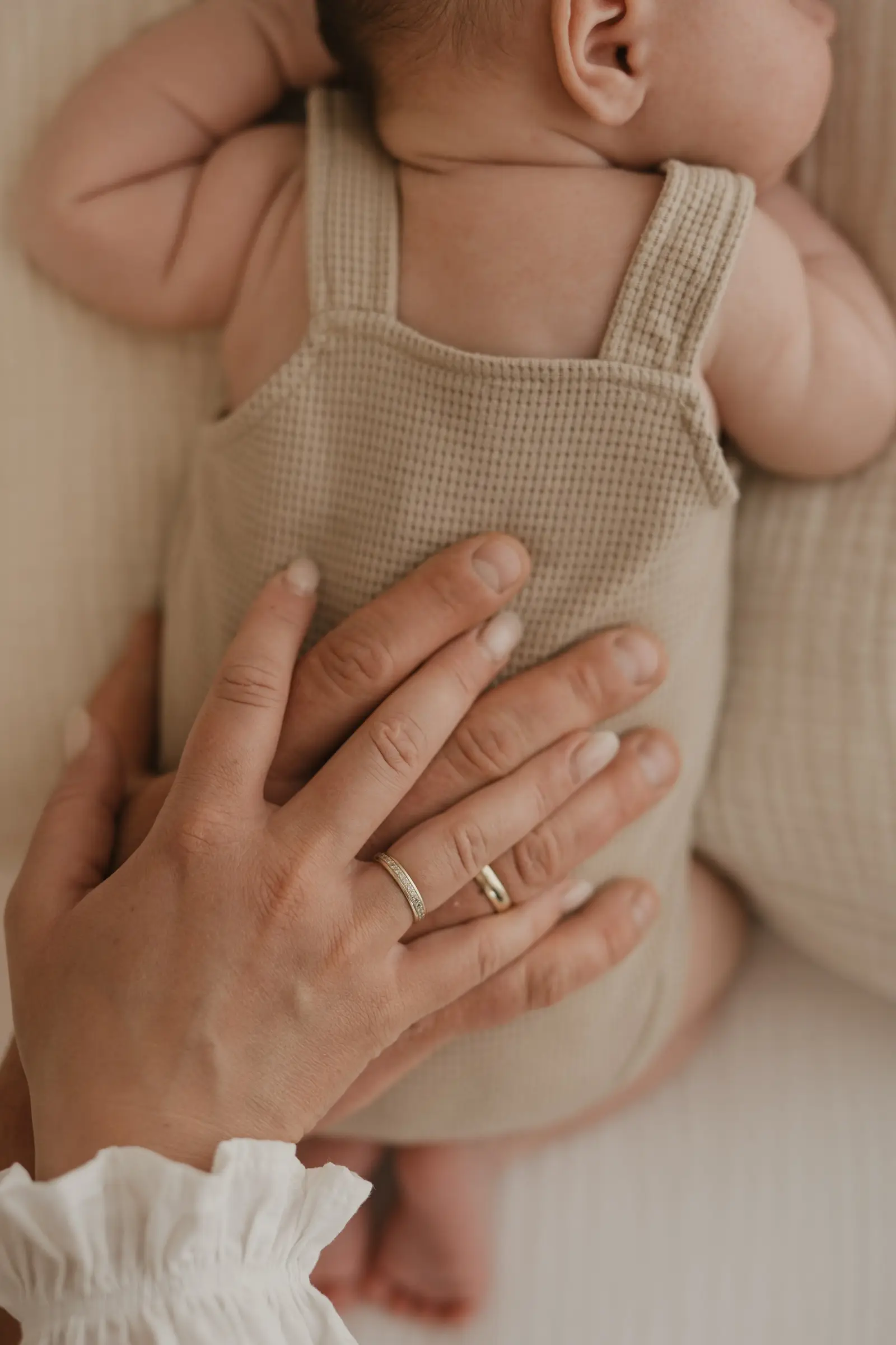 Babybauch im beigen Strampler, Elternhand mit Ehering